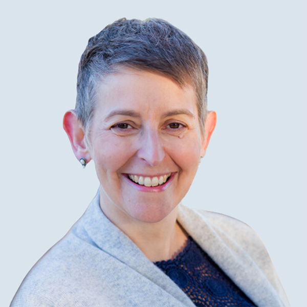 Sarah is a light-skinned woman with short dark hair. She is looking at something out of the shot and laughing. She is wearing a navy blouse and a grey knit cardigan. She is pictured against a blue-grey background.