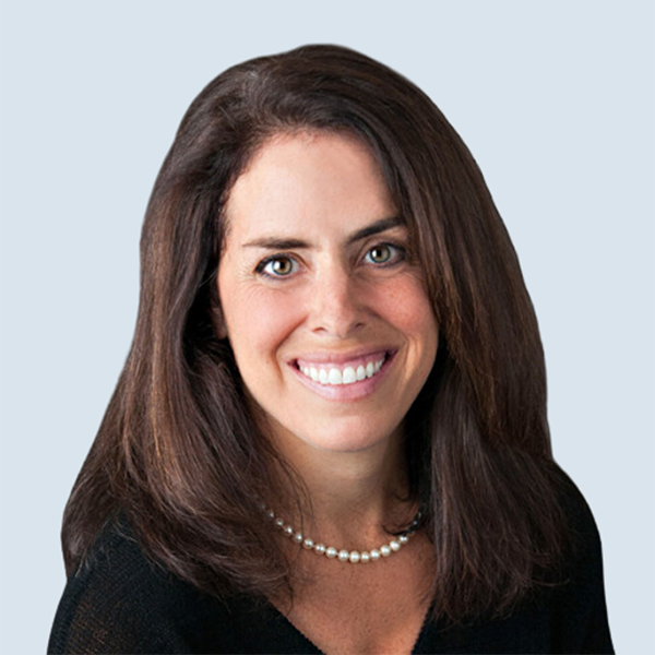 Janet is a light-medium skinned woman with long, dark hair. She is wearing a dark, v-neck sweater and a short strand of white pearls. She is pictured smiling in front of a blue-grey background.