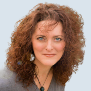 Dawn is a light-skinned woman with bright blue eyes and long, curly hair. She is wearing large silver earrings and a grey shirt. She is pictured smiling broadly against a blue-grey background.