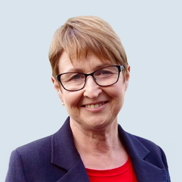 Trisha Carter is a fair skinned woman smiling warmly at the camera. She has light brown hair and dark framed glasses. Trisha is wearing a dark blue jacket over an orange top and is in front of a blue-grey background.