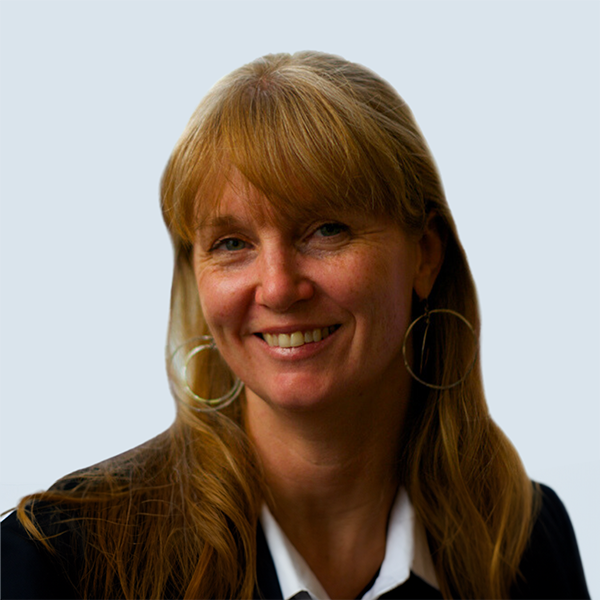 Laurie is a light-skinned woman with freckles. Her hair is long and strawberry-blond, and she has bangs. She is wearing large hoop earrings and a black jacket over a white collared shirt. She is pictured with a relaxed, open smile in front of a blue-grey background.