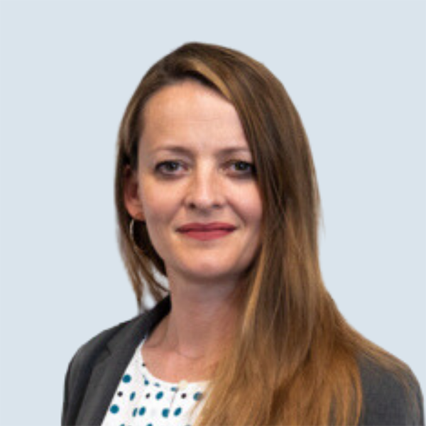 Kasia is a light-skinned woman with green/hazel eyes and long, straight, brownish hair. She is wearing a blue spotted shirt and a grey blazer. She is pictured against a blue-grey background.