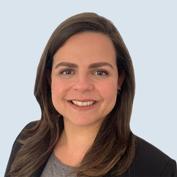 Flor is a medium-skinned woman with big brown eyes and long, wavy brunette hair. She is wearing a black blazer with a grey shirt. She is pictured against a blue-grey background.