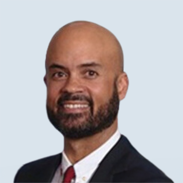 Mark is an African-American man with a dark brown beard and brown eyes. He is wearing a black suit jacket over a white collared shirt and red tie. He is pictured smiling broadly in front of a blue-grey background.