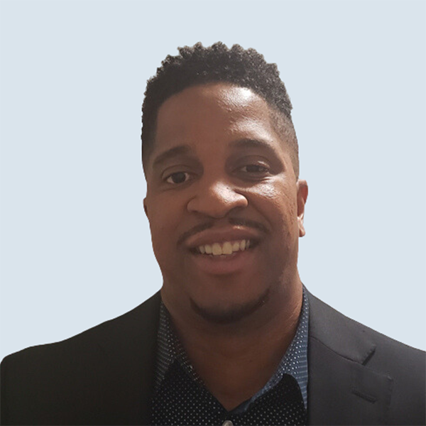 Aarin is a medium-dark skinned man with short dark hair. He is looking towards the camera with his head slightly tilted and smiling. He is wearing a dark blue, patterned collared shirt with a black blazer on top. He is pictured against a blue-grey background.