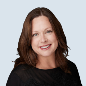 Mary is a light-skinned woman with long dark brown hair and blue eyes. She is wearing a black blouse. She is pictured smiling against a blue-grey background.