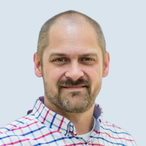 Jerry is a light-skinned man with short, thin, brown hair and facial hair. He is looking directly at the camera and smiling. He is wearing a white shirt with red and blue stripes which are cross-hatched. He is pictured against a blue-grey background.