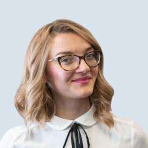 Gradiola is a light/medium-skinned woman with green eyes and long, wavy blonde hair. She is wearing glasses and a white top with a black bow. She is pictured smiling against a grey-blue background.