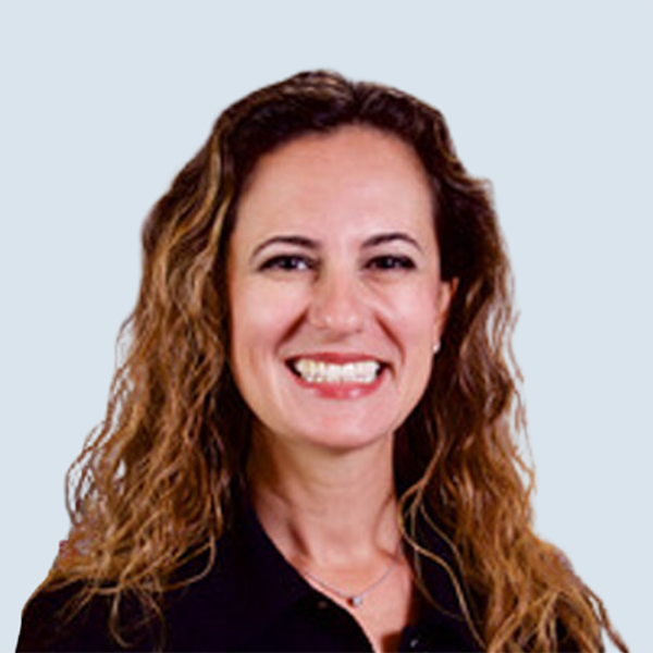 Joanna is a light-skinned woman with brown eyes and wavy brown hair. She is looking at something out of the shot and smiling. She is wearing a black blazer and is pictured against a blue-grey background.