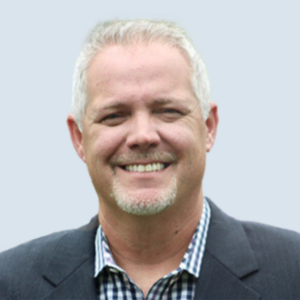 Bill is a light-skinned man with gray hair and a gray goatee. He wears a black blazer over a blue and white checkered shirt. He is pictured with broad smile against a blue-grey background.