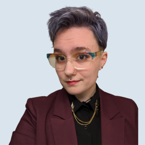 Sarah is a light-skinned person with short teal hair. They are looking at the camera with a small smile. They are wearing a maroon blazer and black button down. They are pictured in front of a blue-grey background.