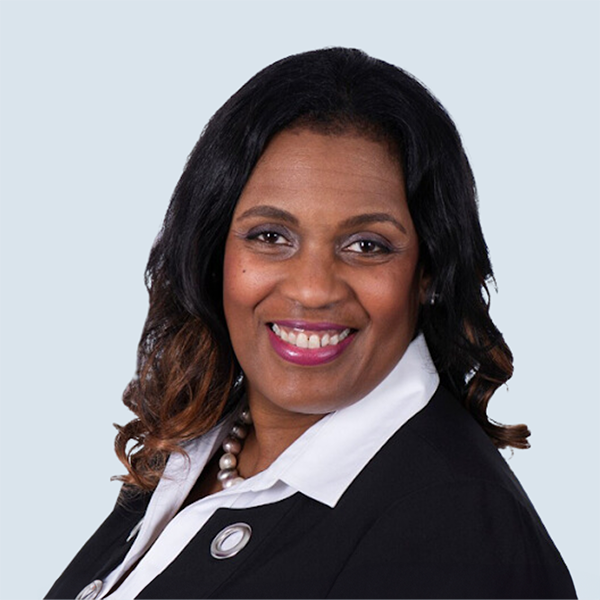 Tayna is a medium-dark skinned female with dark brown eyes and shoulder length dark brown hair. She is wearing a black jacket with a white collared dress shirt and a pearl necklace. She is pictured smiling against a blue-grey background.