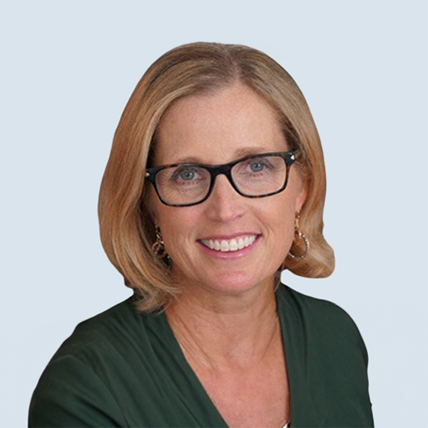 Jodi is a light-skinned woman with shoulder length blonde hair and blue eyes. She is wearing glasses, a dark green blouse and gold earrings. She is pictured smiling broadly against a blue-grey background.