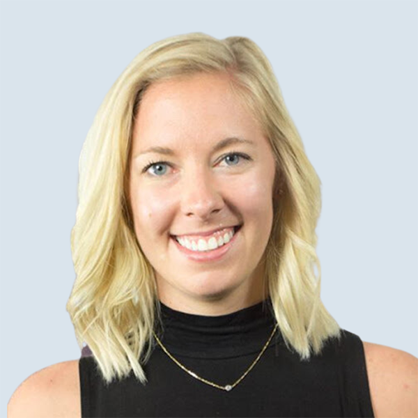 Annie is a light-skinned woman with blue eyes and short, straight blonde hair. She is smiling and wearing a thin gold necklace and a black top. She is pictured against a blue-grey background.