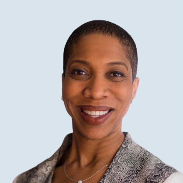 Joyce is a medium-brown skinned woman with brown eyes and short dark brown hair. She has on small studded earrings with a matching pendant necklace. She is wearing a black and white top and is pictured smiling against a blue-grey background.